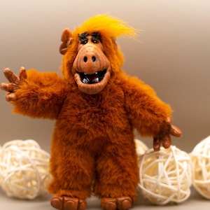 Op de afbeelding: Een bruine, harige pop van het personage Alf, met geel haar en een lachend gezicht. De pop heeft uitgestrekte armen en staat voor witte decoratieve ballen.