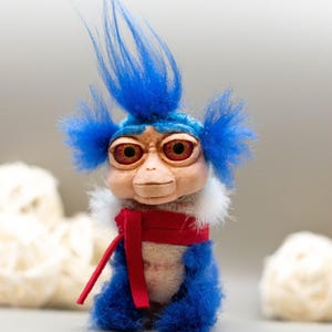 May include: A handmade plush doll with a whimsical design. The doll features a light brown face, large red eyes, and bright blue hair. It wears a red scarf and has blue fur on its body. The doll is a unique, handcrafted item.