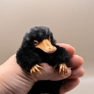 Könnte beinhalten: Ein schwarzes Niffler-Plüschtier mit einem beigen Schnabel und Krallen. Die Kreatur wird in einer Hand gehalten, wobei ihr flauschiges Fell und ihre detaillierten Merkmale hervorgehoben werden. Ein skurriles Sammlerstück.