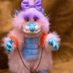 May include: A plush monster doll with pink fur, blue hands and feet, and a blue and white striped belly. It has a purple bow, a chain leash, and a toothy grin. The doll has orange armbands and yellow eyes.