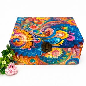 以下が含まれることがあります： 青、オレンジ、黄色、ピンクの色合いで、鮮やかな多色の花柄とペイズリー模様が施された長方形の木箱。箱の前面にはブロンズ調の金属製ラッチが付いており、小さなピンクの花が添えられています。