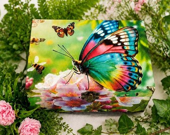 Puede incluir: Una caja rectangular de madera que presenta una mariposa vibrante y multicolor con alas azules, rosas y amarillas posada sobre flores claras. La tapa de la caja también muestra mariposas más pequeñas y abejas sobre un fondo verde suave.