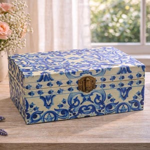 Può includere: Una scatola rettangolare in legno con un motivo decorativo blu e bianco che ricorda le tradizionali piastrelle in ceramica. La scatola è dotata di un coperchio incernierato e di una chiusura centrale in metallo color ottone sul davanti.
