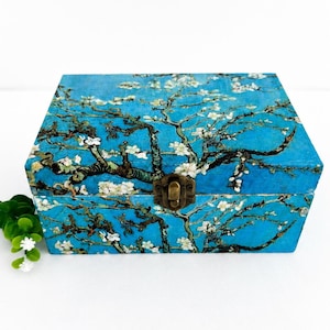 May include: A rectangular wooden box featuring a vibrant blue background with a printed pattern of white almond blossoms on dark brown branches. The box includes a central brass-coloured metal latch on the front and a small green artificial plant.