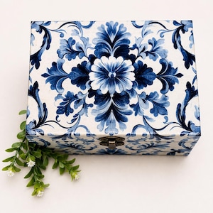 Peut inclure: Une boîte rectangulaire en bois avec une base blanche ornée d'un motif floral bleu symétrique. Le couvercle est fixé par un petit loquet en métal de couleur bronze centré. Un petit brin de feuilles vertes à fleurs blanches est posé à côté.