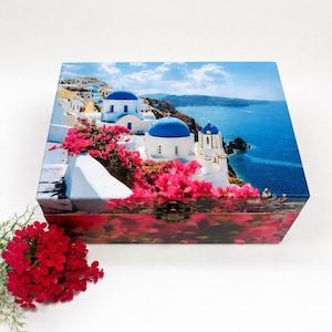 May include: A rectangular wooden keepsake box featuring a printed image of white buildings with blue domes and vibrant pink bougainvillea flowers overlooking the sea. The box has a metal latch on the front and a matching floral print on the side.