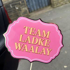 Op de afbeelding: Een roze fotohokjesprop met gouden rand en de woorden "TEAM LADKE WAALAY" in gouden letters. De prop wordt vastgehouden door een houten stok.