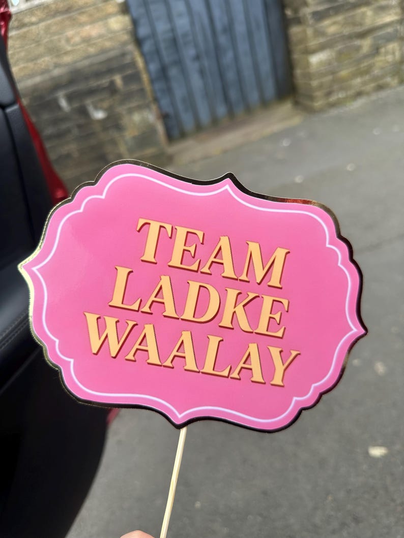 Op de afbeelding: Een roze fotohokjesprop met gouden rand en de woorden "TEAM LADKE WAALAY" in gouden letters. De prop wordt vastgehouden door een houten stok.