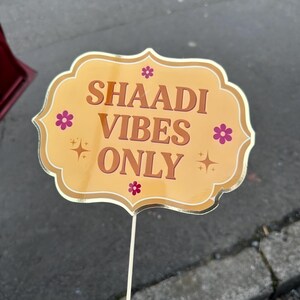 Op de afbeelding: Een fotohokjesprop met een gouden rand en een lichtoranje achtergrond. De prop heeft de woorden "SHAADI VIBES ONLY" in bruine letters, met kleine roze bloemen en steraccenten. De prop wordt vastgehouden door een witte stok.