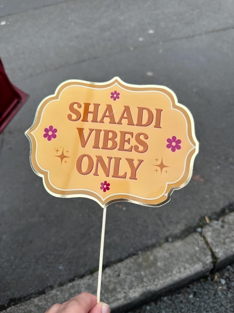 Op de afbeelding: Een fotohokjesprop met een gouden rand en een lichtoranje achtergrond. De prop heeft de woorden "SHAADI VIBES ONLY" in bruine letters, met kleine roze bloemen en steraccenten. De prop wordt vastgehouden door een witte stok.