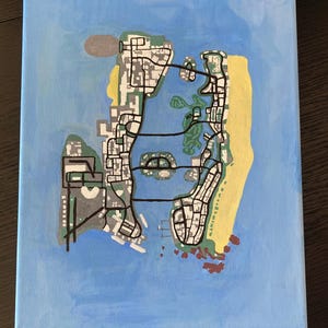 Op de afbeelding: Een handgeschilderd canvas met een kaart van een stad, waarschijnlijk een fictieve locatie. De kaart toont wegen, gebouwen en groene en gele landmassa's omringd door blauw water. Het kunstwerk staat op een rechthoekig canvas.
