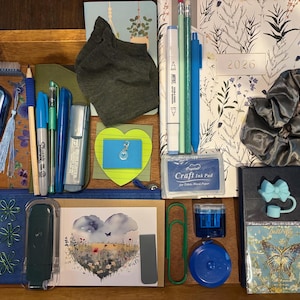 May include: A wooden tray containing a collection of stationery and accessories including a 2026 planner, blue pens, pencils, a grey scrunchie, a heart-shaped sticky note, a craft ink pad, a butterfly-themed card, and various office supplies.