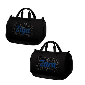 May include: Two black duffel bags with top handles and shoulder straps. One bag features the name "Ziya" in blue script with a dotted burst design, and the other has "Zara" in the same style. Both bags are cylindrical in shape.