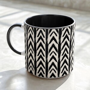 May include: A ceramic mug featuring a black and white chevron pattern with vertical stripes. The exterior displays a repeating hand-painted style arrow design in black against a white background, while the interior and the handle are solid black.