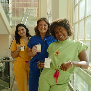 May include: Three people wearing medical scrubs in yellow, blue, and green. Each person holds a small rectangular card. The background includes a staircase and large windows.