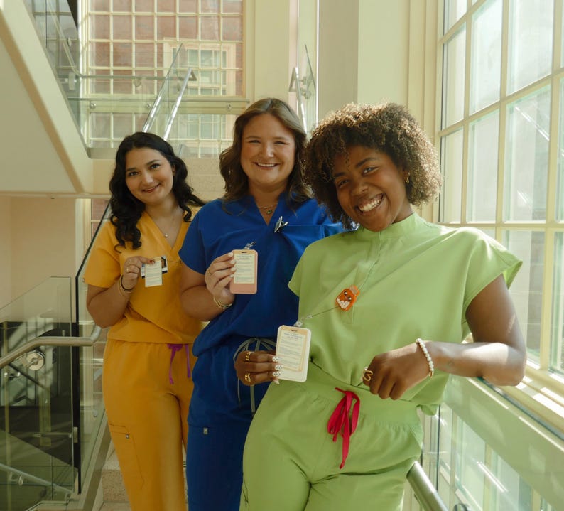 May include: Three people wearing medical scrubs in yellow, blue, and green. Each person holds a small rectangular card. The background includes a staircase and large windows.