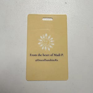 May include: A rectangular, pale yellow card with a hole at the top. It features a white smiling sun graphic and the text "From the heart of Madi P. @DoseofSunshineRx".