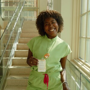 May include: A person wearing a light green scrub set with a matching top and trousers. The top has short sleeves and a mandarin collar. A lanyard with a cartoon character charm and a name tag hangs from the neck. The trousers have a red drawstring.