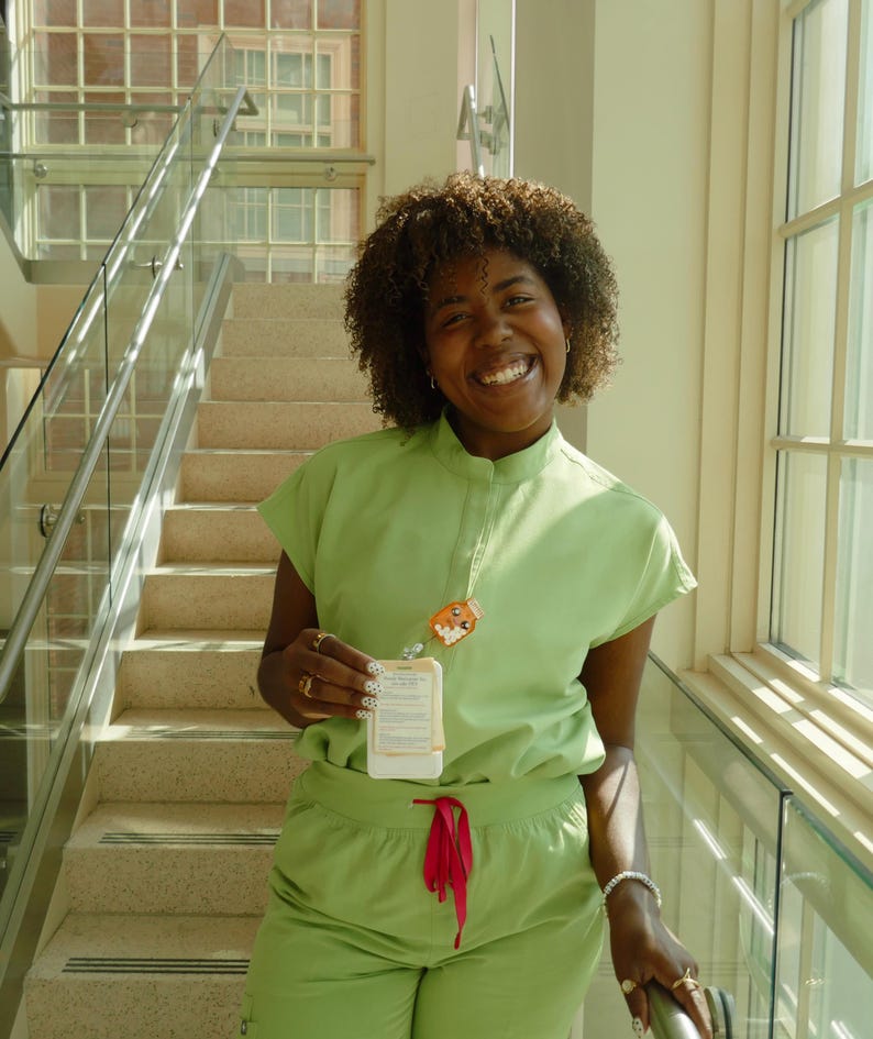 May include: A person wearing a light green scrub set with a matching top and trousers. The top has short sleeves and a mandarin collar. A lanyard with a cartoon character charm and a name tag hangs from the neck. The trousers have a red drawstring.