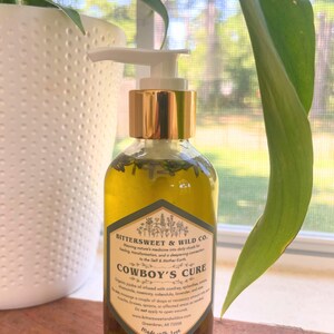 May include: A clear glass bottle of "Cowboy's Cure" oil with a gold pump top. The label features text and botanical illustrations. The bottle is filled with a yellow liquid and green herbs. The background includes a white pot and green leaves.