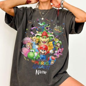 Puede incluir: Camiseta gris oscuro con un gr&aacute;fico colorido de Mario y otros personajes del juego Super Mario Galaxy. El dise&ntilde;o incluye el texto "THE SUPER MARIO GALAXY" y un espacio para un nombre. La camiseta tiene mangas cortas.