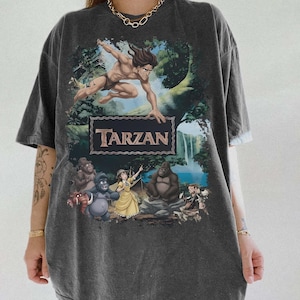 May include: Dark gray t-shirt featuring a Tarzan graphic. The design includes Tarzan swinging through a jungle scene, the word "Tarzan" in a brown frame, and various cartoon characters. The shirt has short sleeves and a relaxed fit.