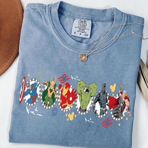 Puede incluir: Una camiseta azul claro con un gr&aacute;fico de varios superh&eacute;roes en fila. La camiseta tiene cuello redondo y mangas cortas. Un collar dorado con un colgante est&aacute; drapeado sobre la camiseta. La camiseta est&aacute; etiquetada como "Comfort Colors".