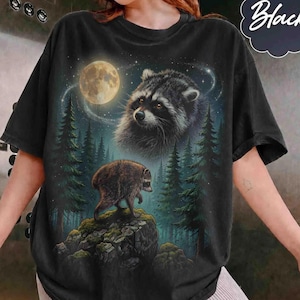 May include: A black t-shirt featuring a graphic print of a raccoon standing on a rocky outcrop in a forest at night. Above the raccoon, a larger raccoon face appears in the sky near a full moon. The scene includes tall pine trees and stars.