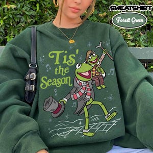 May include: A forest green crewneck sweatshirt featuring a graphic of a green frog wearing a scarf and top hat, holding a cane, with the text 'Tis' the Season' in light green. A smaller frog sits on the larger frog's shoulder.
