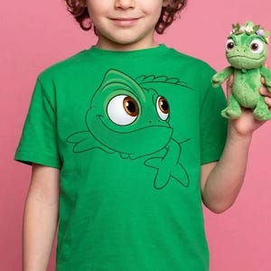 May include: A bright green short-sleeved t-shirt featuring a large graphic print of a cartoon chameleon with wide brown eyes. The shirt is paired with a small, plush green chameleon toy that has a crown of tiny colorful flowers on its head.