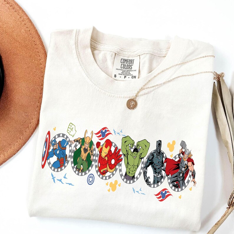 Puede incluir: Camiseta de color crema con un gr&aacute;fico colorido de varios superh&eacute;roes de Marvel en marcos ovalados. La camiseta tiene cuello redondo y mangas cortas. Un collar dorado con un colgante descansa sobre la camiseta. La camiseta tiene una etiqueta que dice "Comfort Colors".