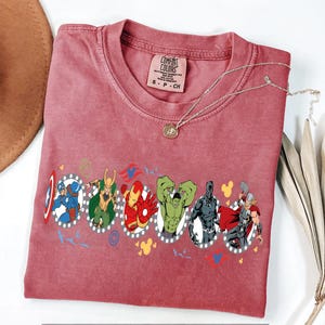 Puede incluir: Una camiseta Comfort Colors color granate con un gr&aacute;fico de superh&eacute;roes en marcos ovalados. La camiseta tiene cuello redondo y mangas cortas. Un collar plateado con un colgante est&aacute; drapeado sobre la camiseta.