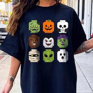 May include: A dark navy blue t-shirt featuring a graphic print of nine square-shaped toy heads arranged in a grid. The heads depict various Halloween characters including a zombie, pumpkin, skull, werewolf, vampire, witch, mummy, alien, and ghost.