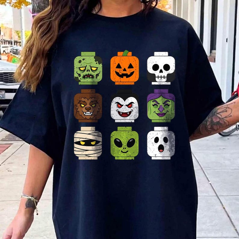 May include: A dark navy blue t-shirt featuring a graphic print of nine square-shaped toy heads arranged in a grid. The heads depict various Halloween characters including a zombie, pumpkin, skull, werewolf, vampire, witch, mummy, alien, and ghost.