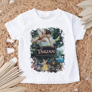 May include: White t-shirt featuring a colorful graphic of Tarzan jumping, with the word "Tarzan" on a sign. The design includes jungle elements and various characters from the animated movie.