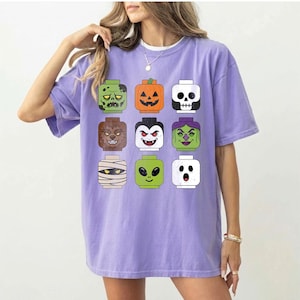 May include: A light purple short-sleeve t-shirt featuring a graphic print of nine stylized toy-like character heads arranged in a grid. The characters include a zombie, jack-o'-lantern, skull, werewolf, vampire, witch, mummy, alien, and ghost.