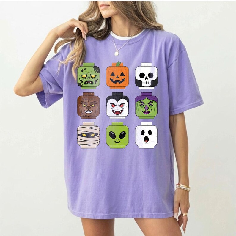 May include: A light purple short-sleeve t-shirt featuring a graphic print of nine stylized toy-like character heads arranged in a grid. The characters include a zombie, jack-o'-lantern, skull, werewolf, vampire, witch, mummy, alien, and ghost.