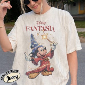 May include: An ivory short-sleeve t-shirt featuring a graphic of Mickey Mouse as the Sorcerer's Apprentice from Disney's Fantasia. Mickey wears a blue star-patterned hat and a red robe, holding a glowing wand. The text Disney Fantasia is printed above.