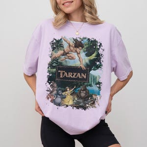 May include: A light purple t-shirt featuring a graphic of Tarzan and other characters from the animated movie. The word "Tarzan" is displayed in a brown, vintage-style font. The shirt has short sleeves and a relaxed fit.