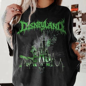 Puede incluir: Camiseta negra con un gr&aacute;fico verde que dice "Disneyland" en una fuente puntiaguda. El gr&aacute;fico presenta una silueta verde de un castillo con fuegos artificiales en el fondo. La camiseta es de color oscuro.