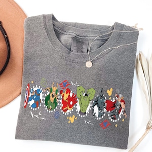Puede incluir: Camiseta gris Comfort Colors con un gr&aacute;fico colorido de varios superh&eacute;roes. El dise&ntilde;o incluye al Capit&aacute;n Am&eacute;rica, Loki, Iron Man, Hulk, Pantera Negra y Thor. Un collar plateado con un colgante redondo descansa sobre la camiseta.