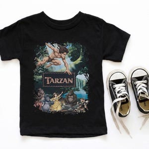 May include: Black t-shirt featuring a colorful graphic of Tarzan, the jungle hero, with the word "Tarzan" in a brown frame. The shirt is paired with black canvas sneakers with white laces.