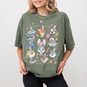 Op de afbeelding: Olijfgroen T-shirt met kleurrijke cartoonkarakterportretten met feesthoeden. Het ontwerp bevat een kikker, een vos, een luiaard, een buffel, een konijn, een schaap, een cheetah, een muis en een kat. Tekst bevat karakternamen.