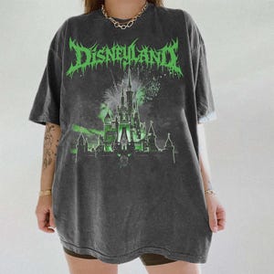 Puede incluir: Camiseta gris oscuro con aspecto desgastado. La parte delantera presenta la palabra "Disneyland" en verde, con letras g&oacute;ticas, sobre una ilustraci&oacute;n de castillo iluminado en verde con fuegos artificiales.