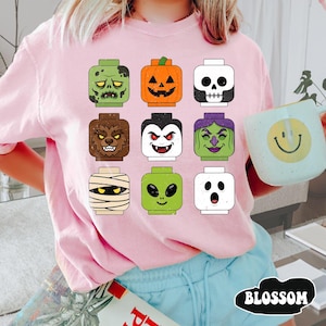 May include: A light pink t-shirt featuring a graphic print of nine stylized toy block heads representing Halloween characters including a zombie, pumpkin, skull, werewolf, vampire, witch, mummy, alien, and ghost, arranged in a three-by-three grid.