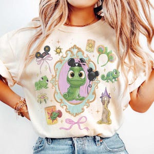 May include: A cream-coloured t-shirt featuring a central illustration of a green chameleon inside an ornate light blue frame. The design includes smaller graphics of a tower, mouse ears, a lantern, flowers, and a pink ribbon, all in a cartoon style.