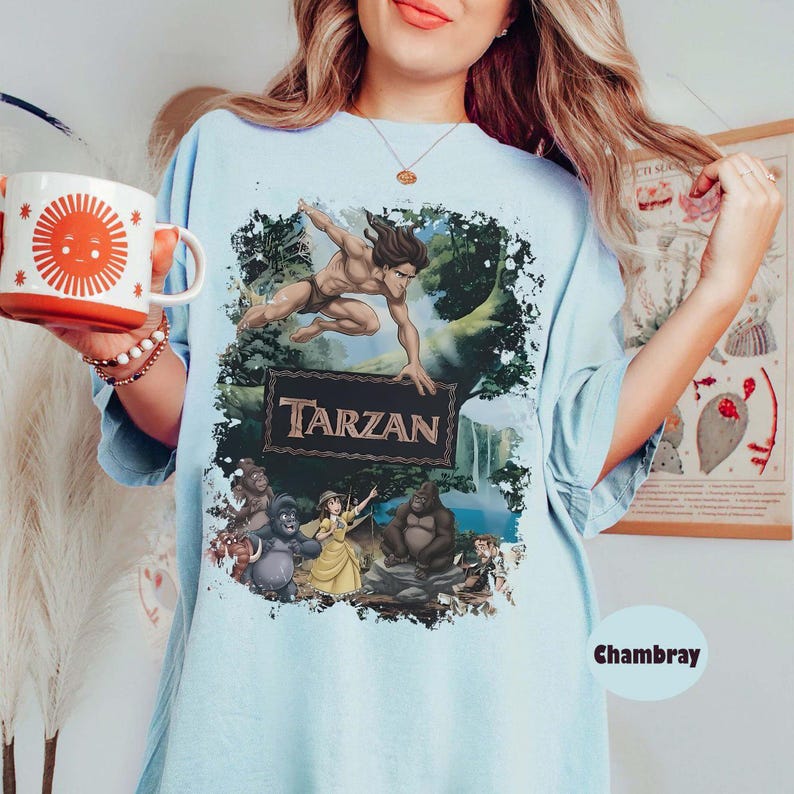 May include: Light blue t-shirt featuring a Tarzan graphic. The design includes Tarzan, jungle scenery, and the word "Tarzan" in a brown font. The shirt has a relaxed fit and is made of a soft material. The shirt is a Chambray color.