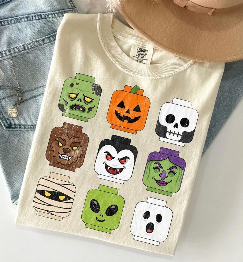 May include: A cream-colored t-shirt featuring a graphic print of nine stylized toy block heads representing Halloween characters, including a zombie, pumpkin, skeleton, werewolf, vampire, witch, mummy, alien, and ghost, with a Comfort Colors label.
