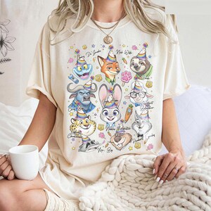 Op de afbeelding: Cr&egrave;mekleurig T-shirt met kleurrijke cartoonfiguren uit Zootopia, elk met een feesthoedje. Het ontwerp bevat Judy Hopps, Nick Wilde en andere personages, met hun namen erboven.
