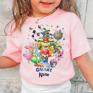 Puede incluir: Una camiseta rosa claro con un gr&aacute;fico colorido de personajes del videojuego "The Super Mario Galaxy". El dise&ntilde;o incluye a Mario, Bowser, la Princesa Peach y otros personajes. Tambi&eacute;n se ve el texto "THE SUPER MARIO GALAXY" y "Name".